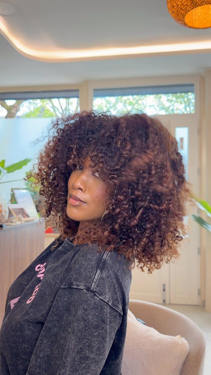 Layered dark curls frame a seated person's face in a bright salon.
