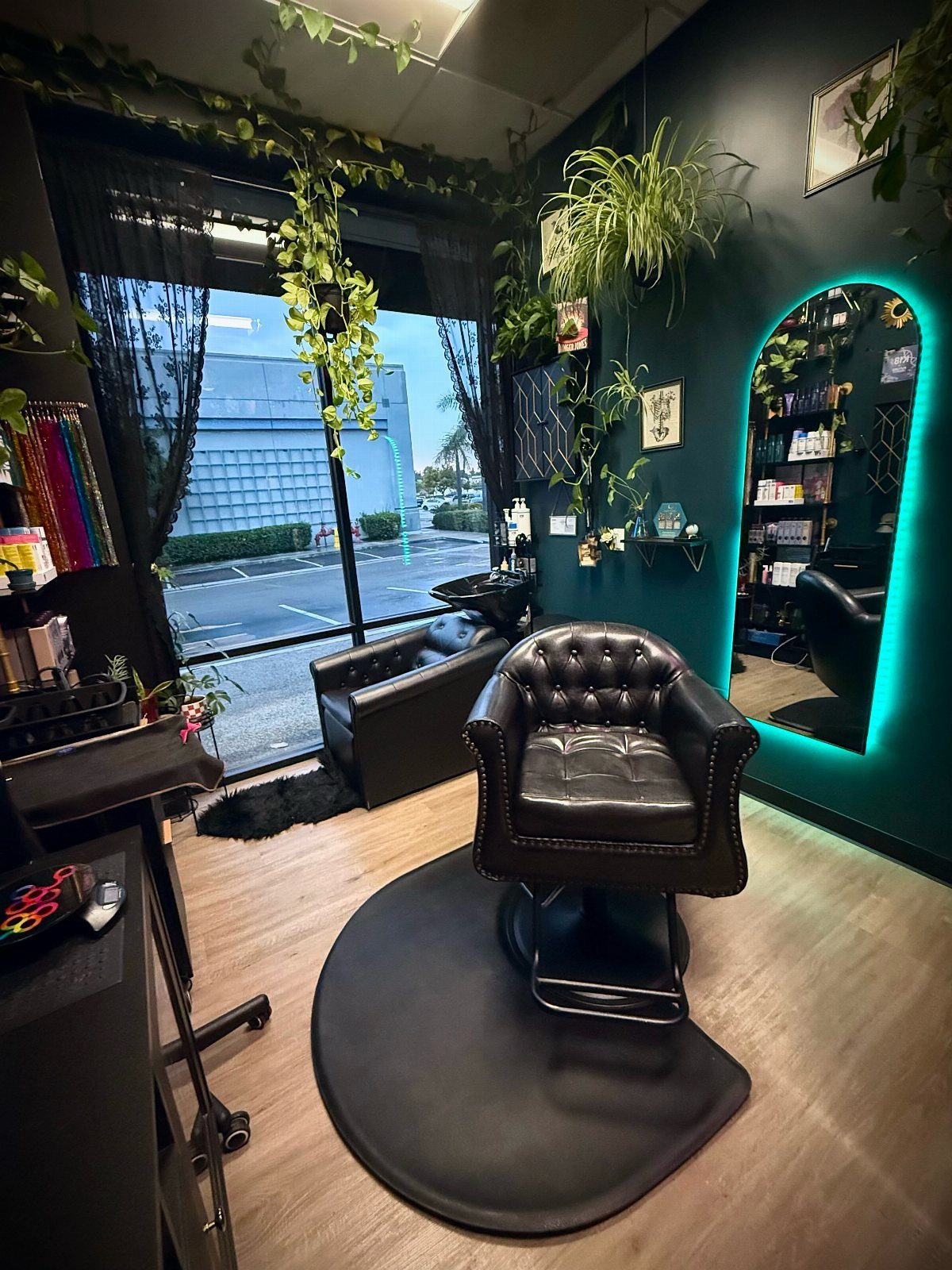 Dark teal salon with leather styling chair, glowing mirror, wood floor, and hanging plants
