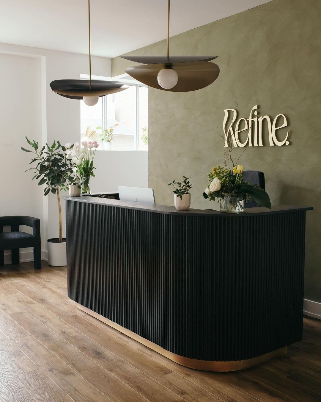 Curved dark wood reception desk beneath pendant lights in a bright minimalist salon