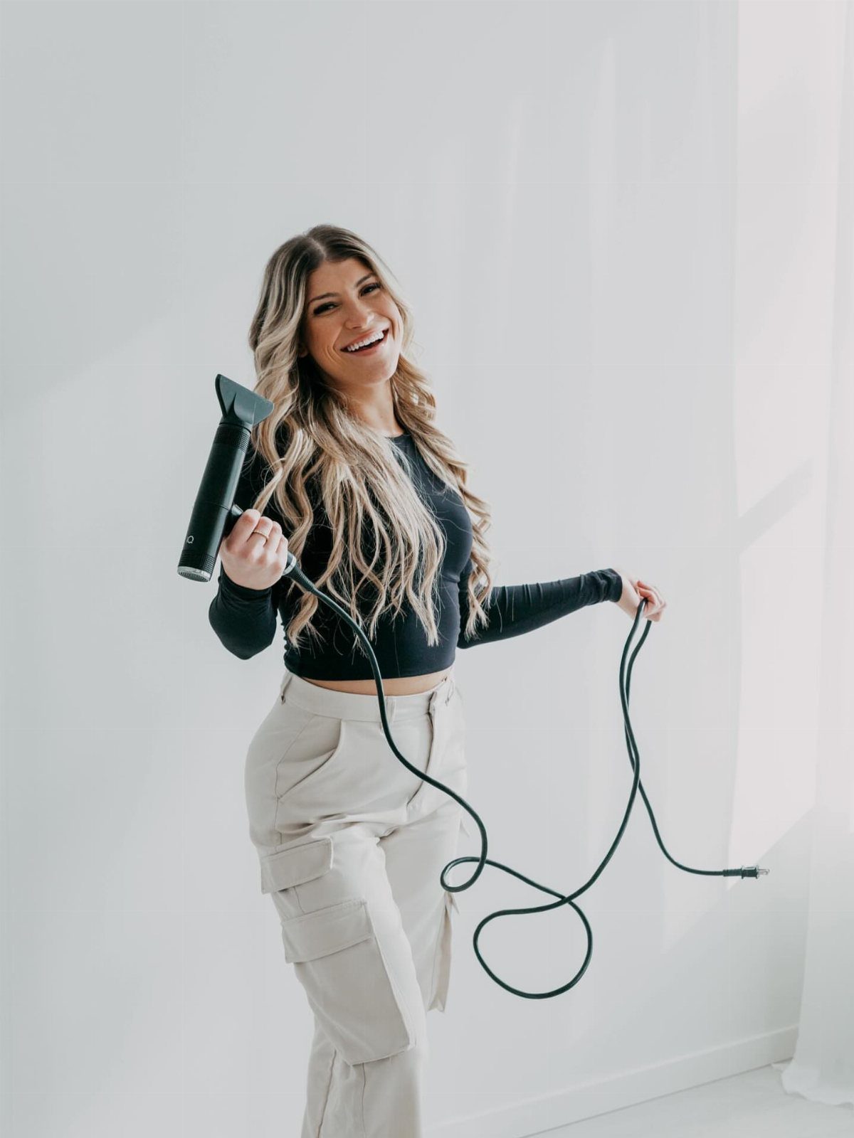 Smiling stylist poses with a black hair dryer in a bright white room