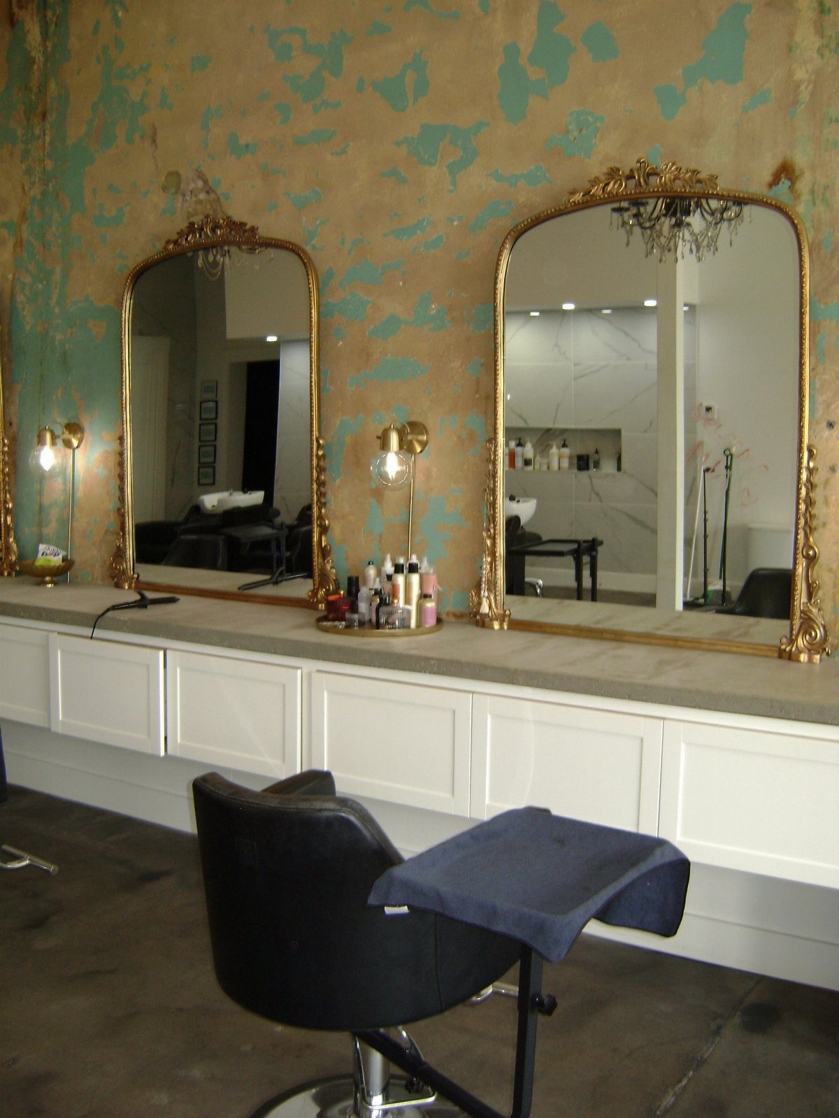 Salon station with ornate mirrors, distressed teal wall, and a black styling chair