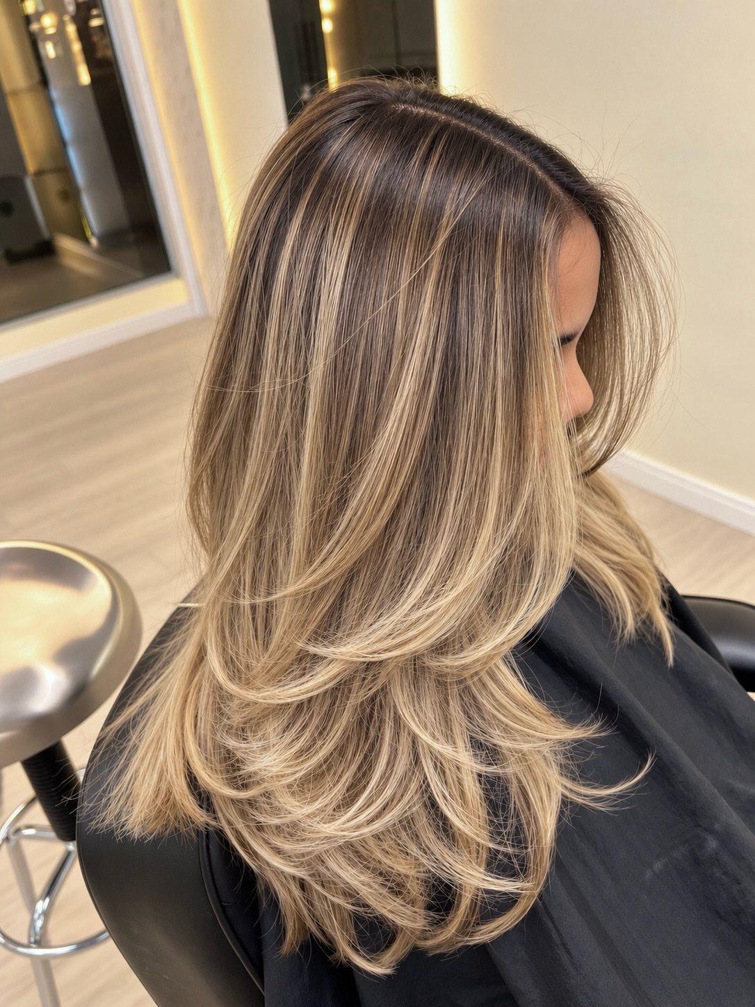 Woman with layered golden ash blonde hair and dark roots in a salon.