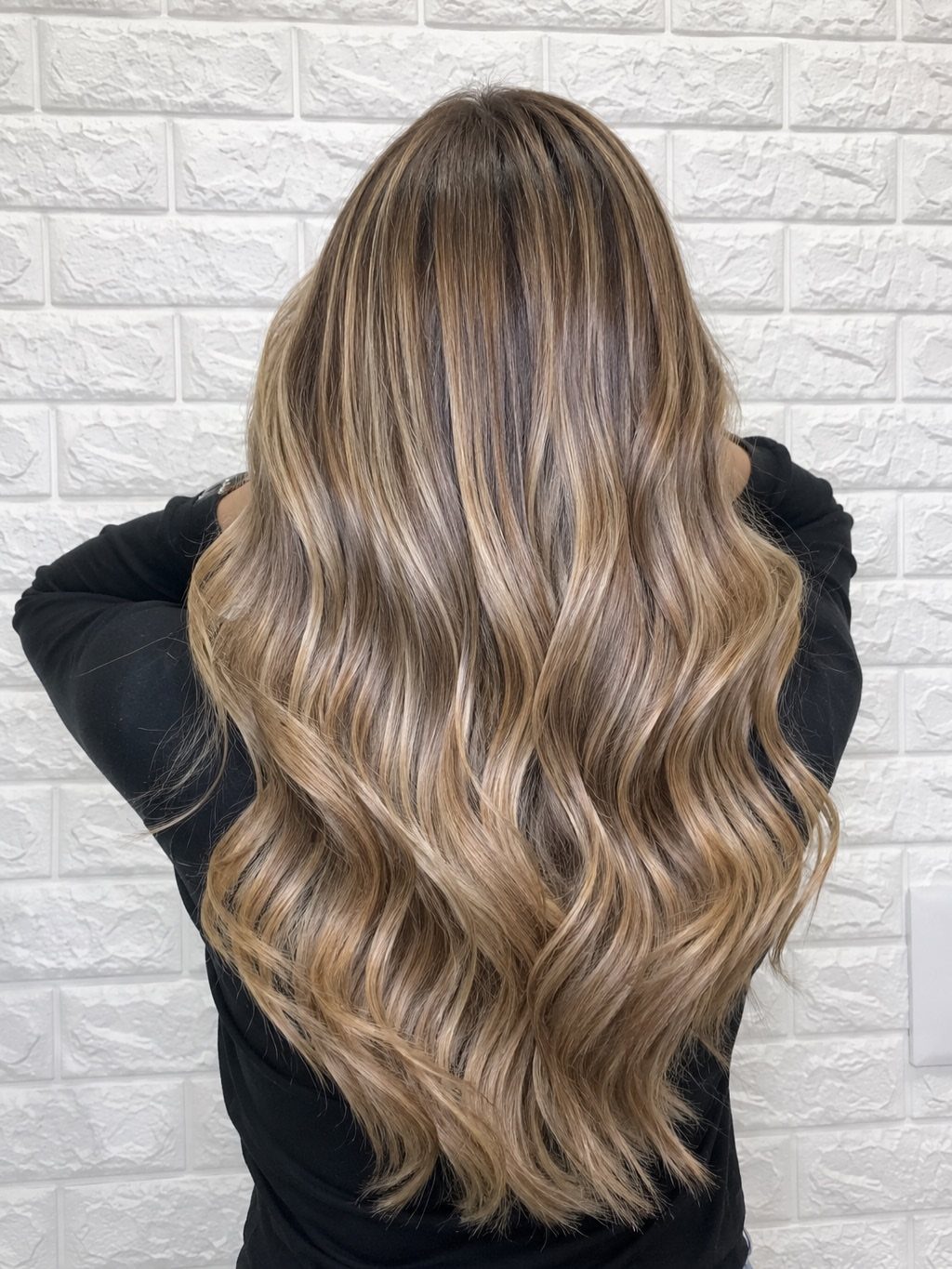 Long wavy honey ash blonde hair with dark roots against a white brick wall.