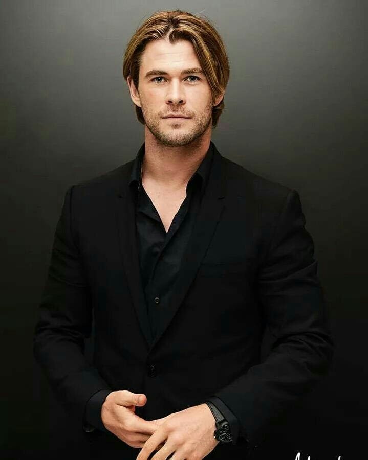 Man with center-parted blond hair wearing a black suit against gray