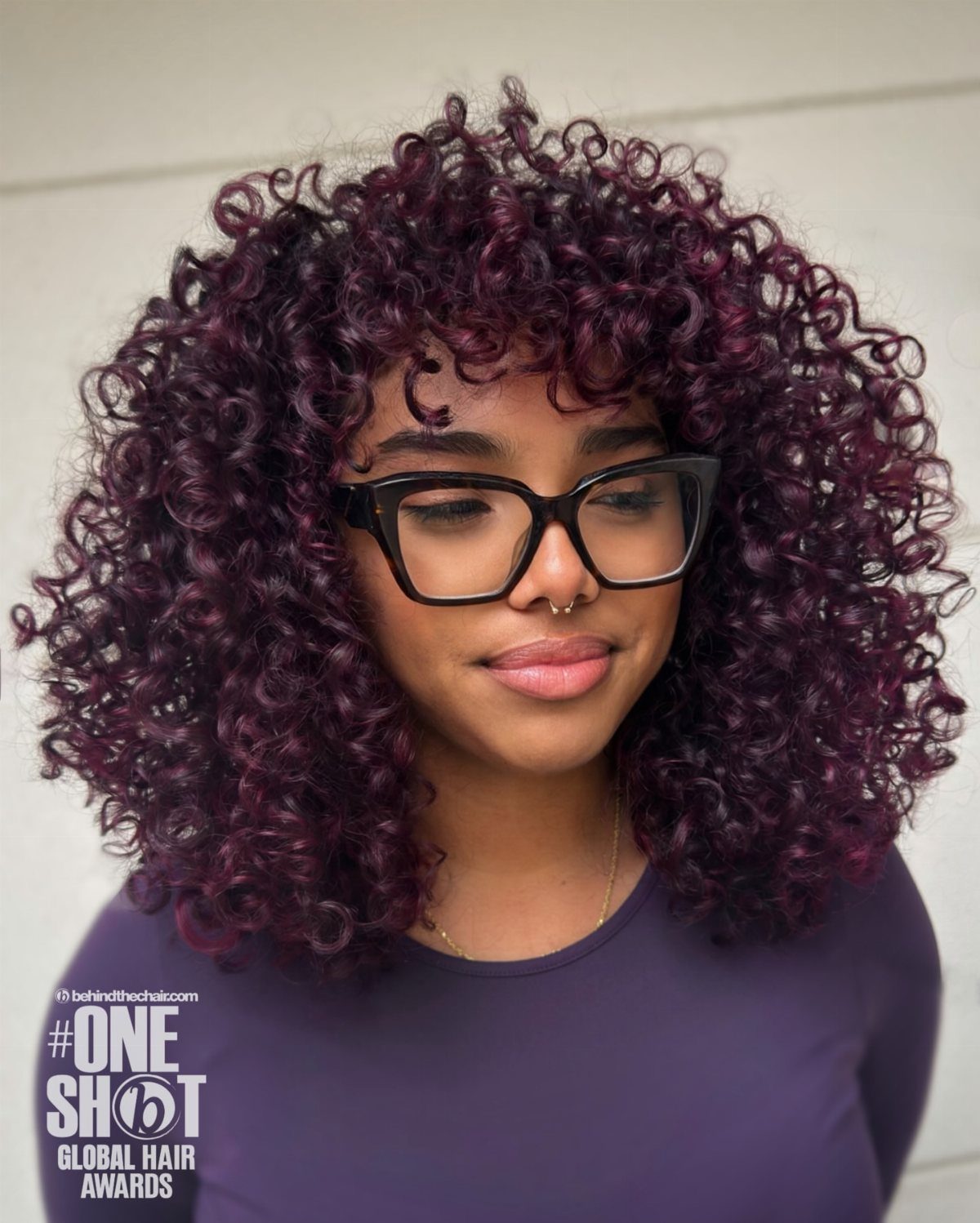 Voluminous dark plum-purple curls frame a person wearing black glasses and a purple top.