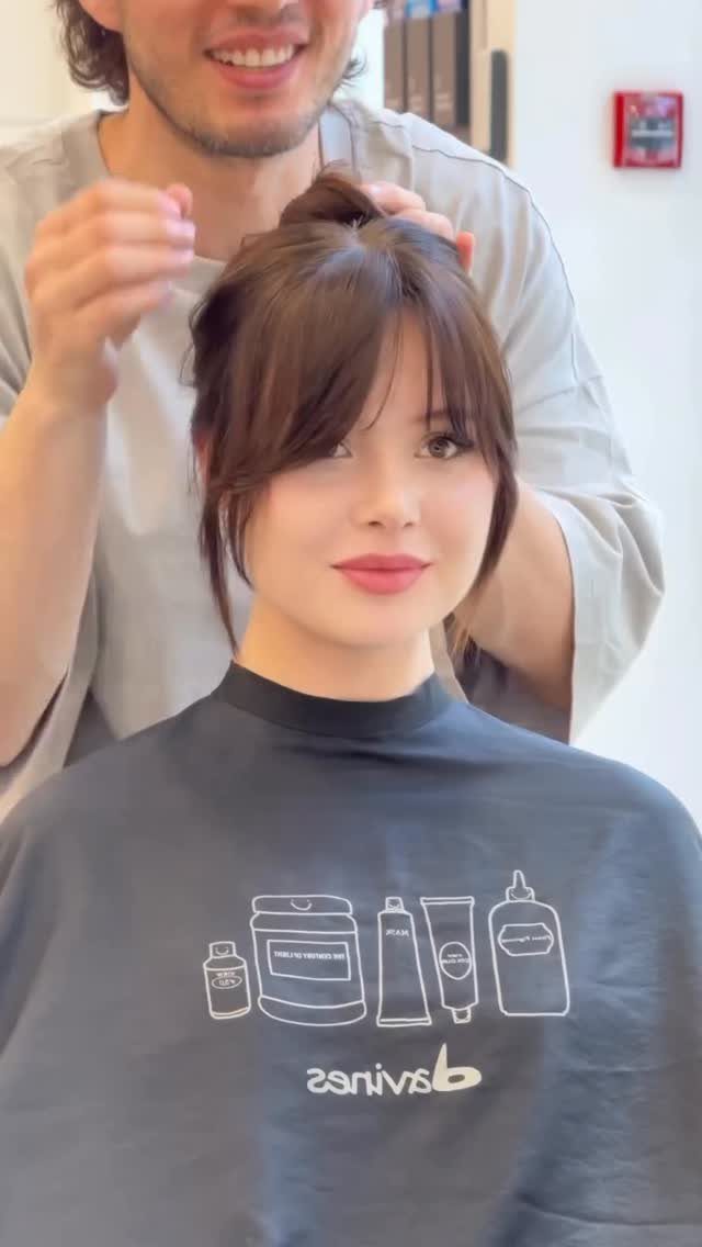 Woman with brown curtain bangs sits in a salon as a stylist arranges her hair