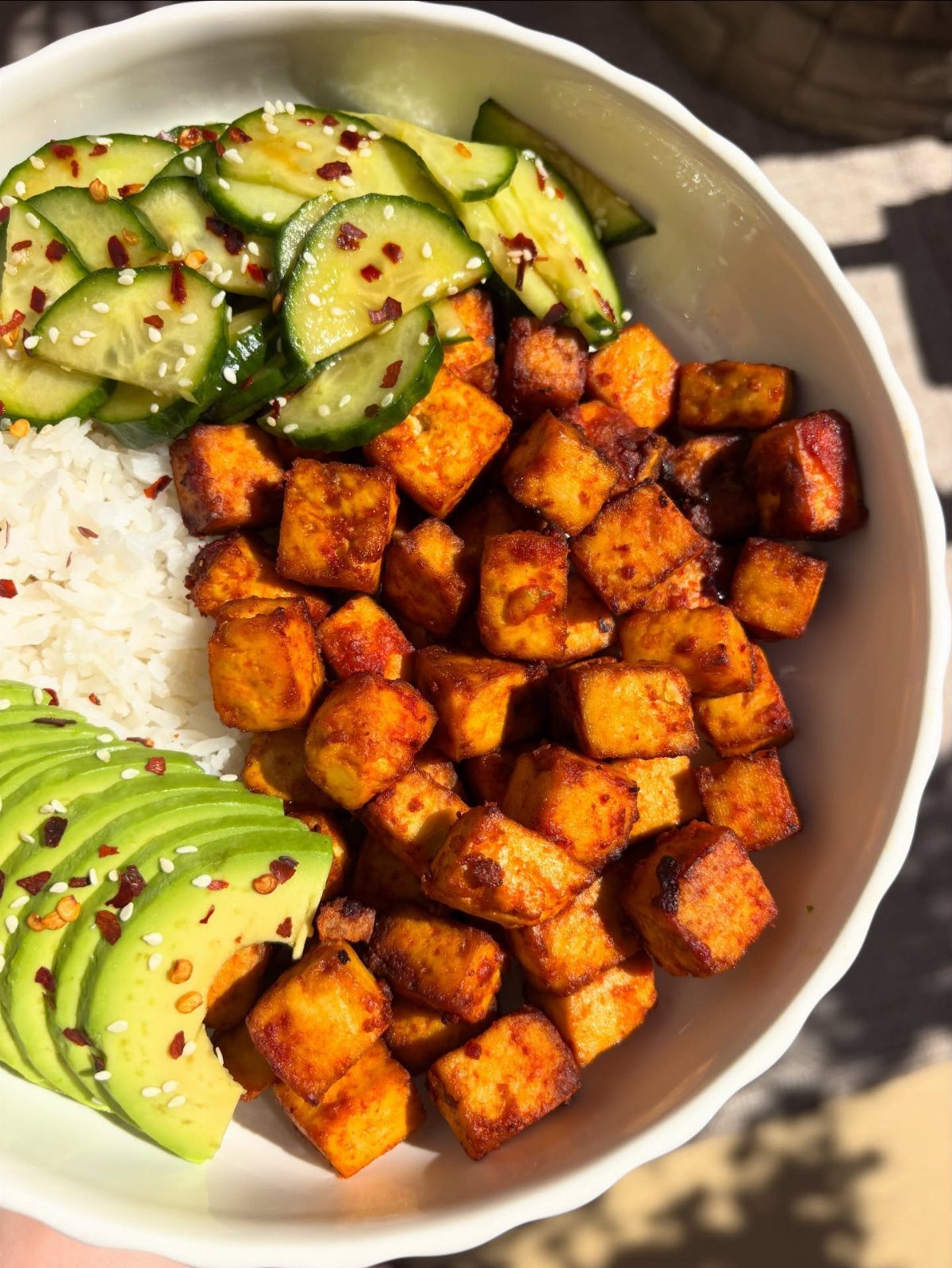 Rice bowl topped with tofu, avocado, cucumber salad, seeds, and red pepper flakes