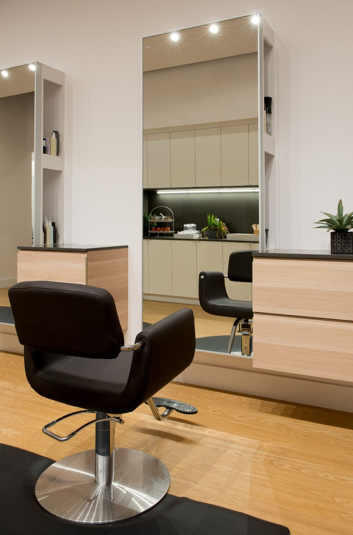 Illuminated salon mirror above light wood cabinets beside a black styling chair