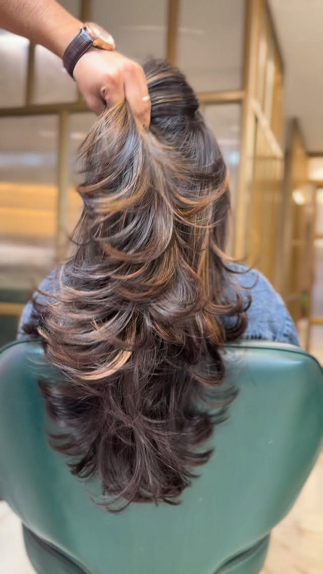 Stylist lifts long brunette balayage waves behind a seated salon client