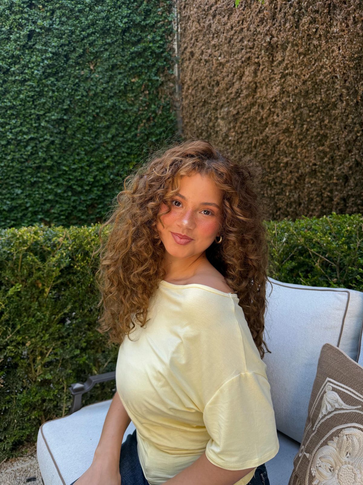 Woman with voluminous brown curls framing her face, seated outdoors in a white top