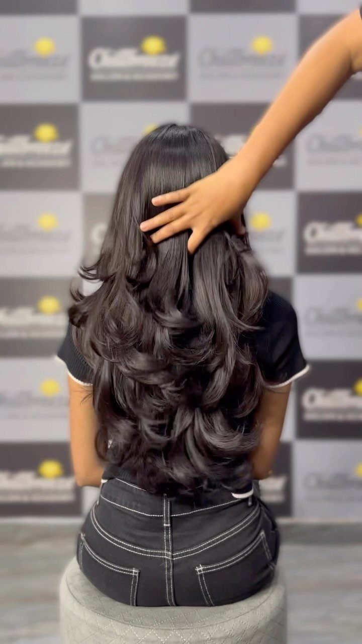 Long dark layered hair falls in glossy curls down a seated person’s back