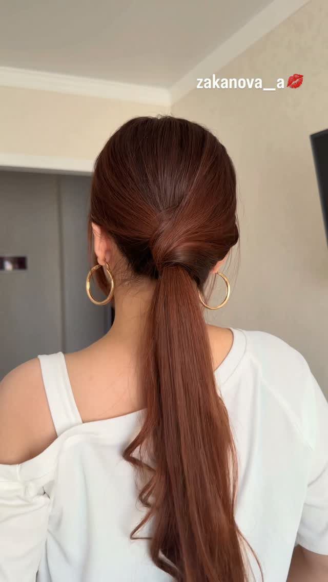 A woman displays a twisted low ponytail with long auburn hair and gold hoops