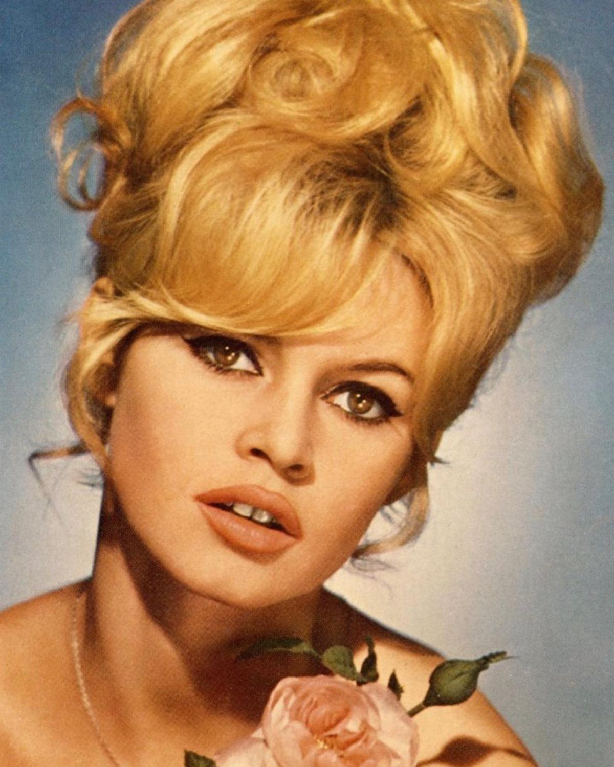 Woman with voluminous curled blonde bouffant hair and a pink rose