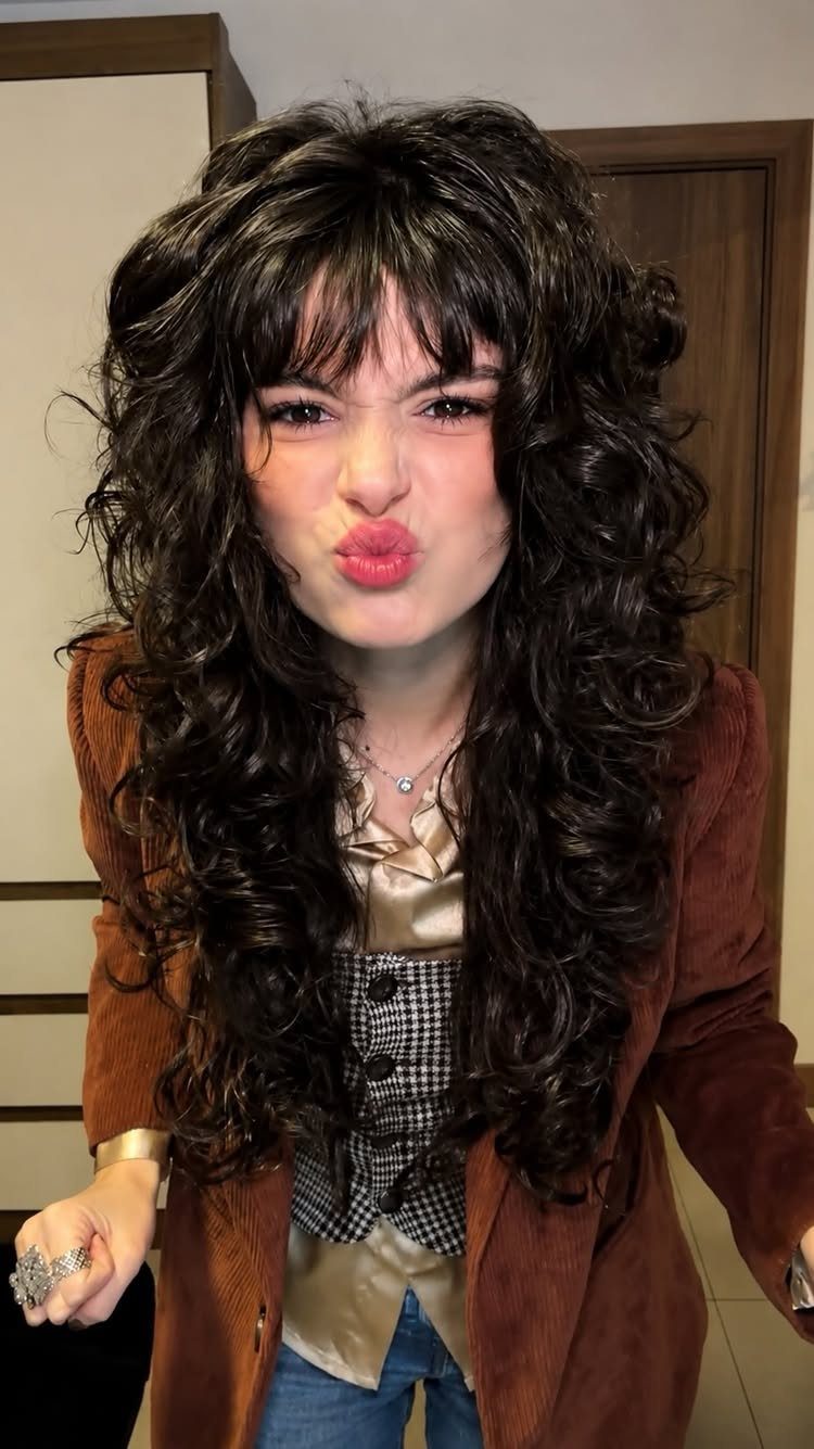 Person posing with long dark curls and curly bangs in a brown jacket