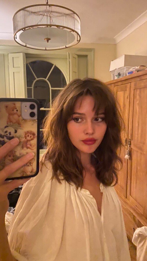 Woman with long wavy brown hair and wispy, side-lengthening bangs takes a mirror selfie