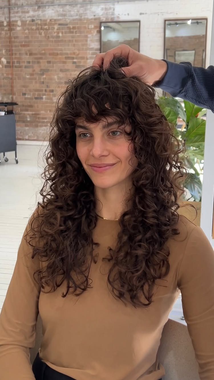 Woman with long dark curls and bangs seated in a salon while a stylist adjusts her hair