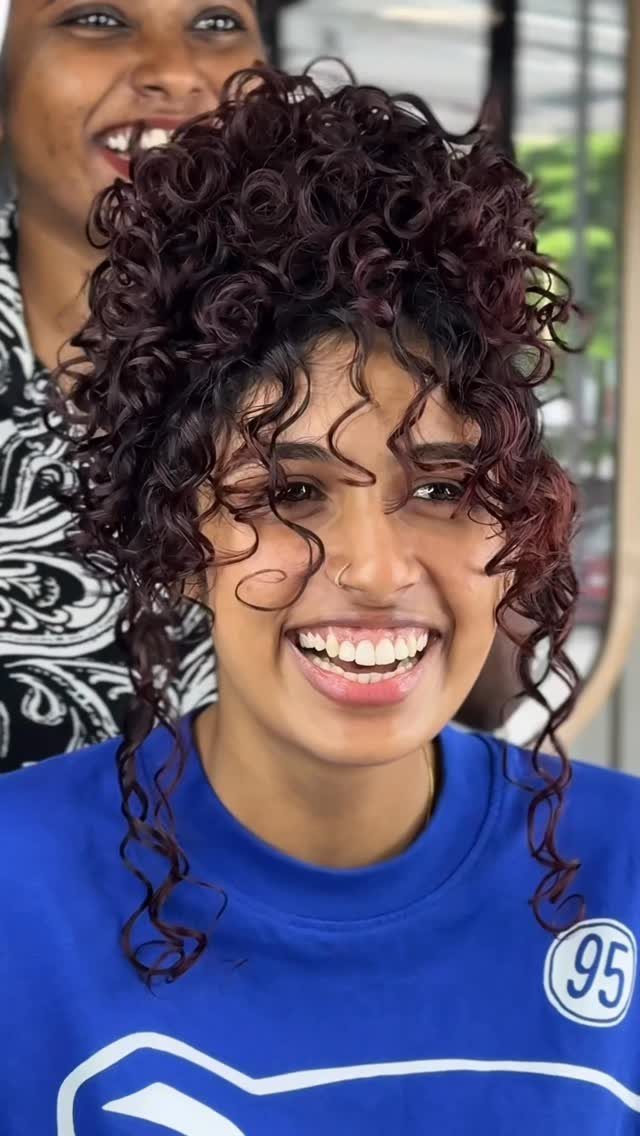 Dark burgundy curls frame a smiling woman's face, with another woman behind her.