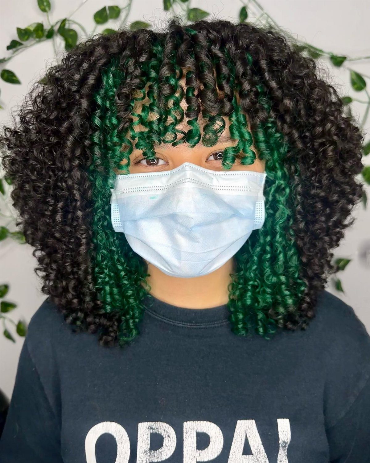 Shoulder-length black and green curls frame a masked person's face
