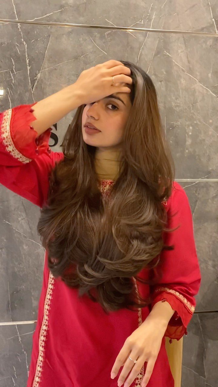 Long layered brown hair styled in loose, voluminous curls against a gray tiled wall