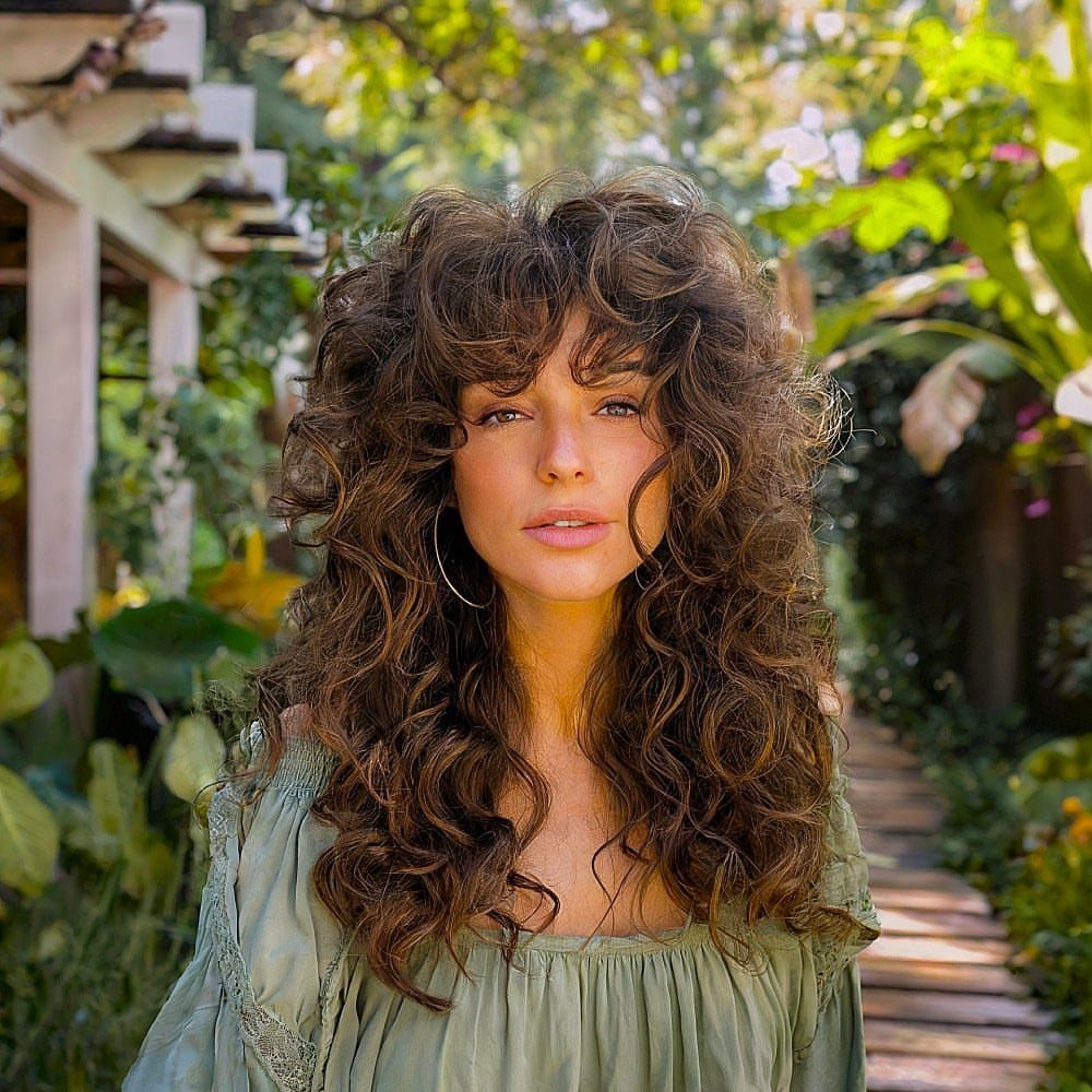 Woman with voluminous dark curls and bangs posing among leafy garden plants