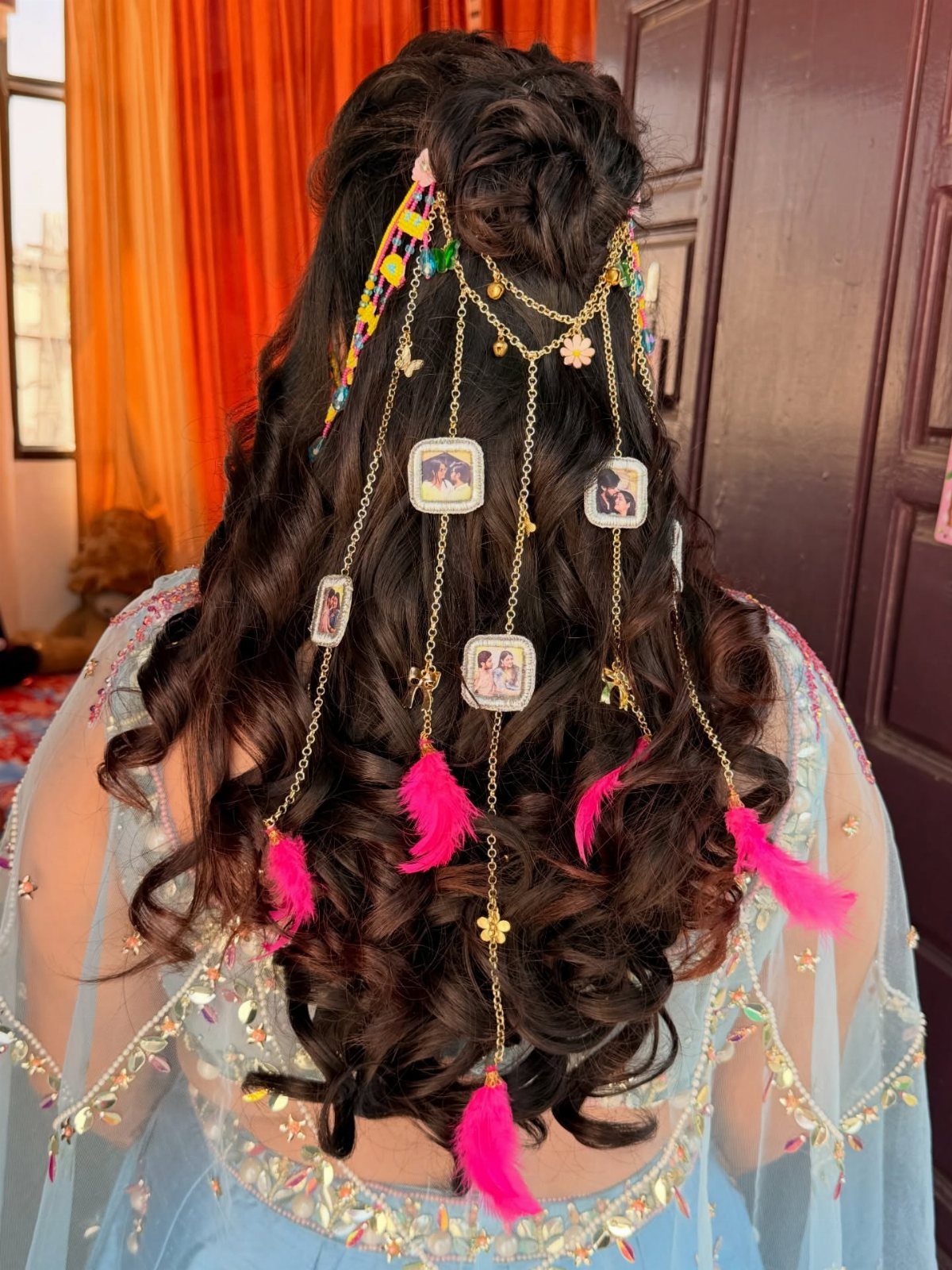 Long curled half-up hairstyle decorated with gold chains, ribbons, ornaments, and pink feathers
