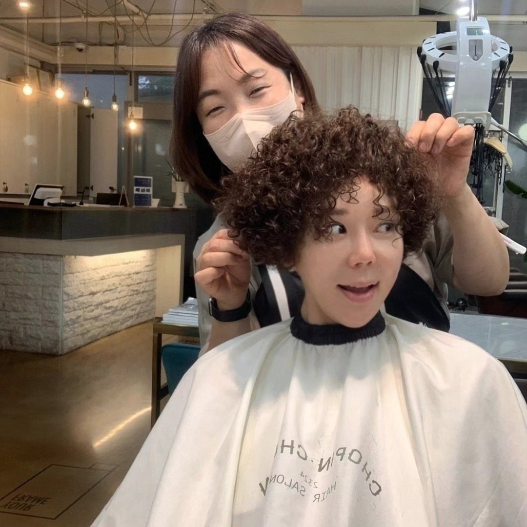 Short brown curls frame a seated person's face as a stylist lifts them in a salon