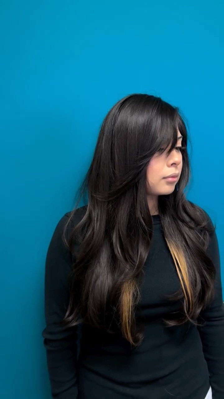 Woman with long dark layered hair and side-swept bangs against a blue wall