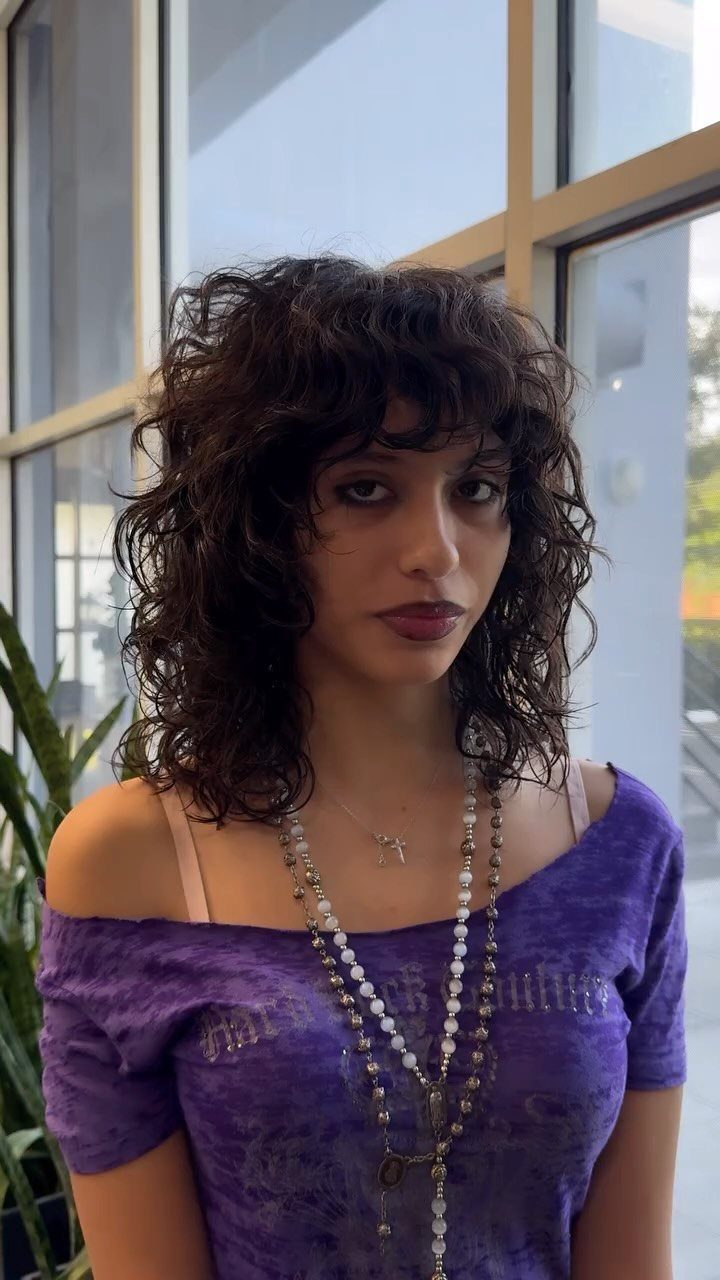 Person with dark curly wolf-cut hair and bangs wearing a purple top