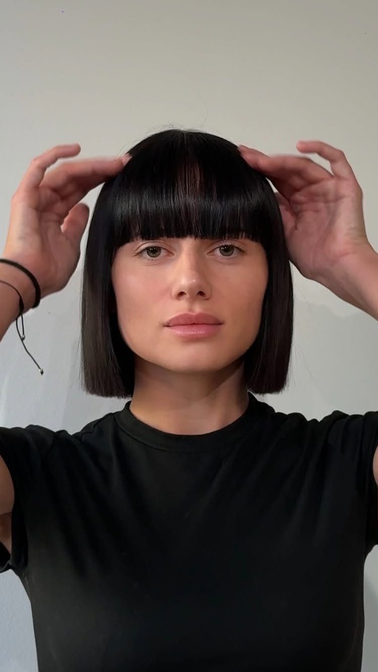 Person adjusts a sleek dark bob with blunt bangs, cut neatly at jaw level.