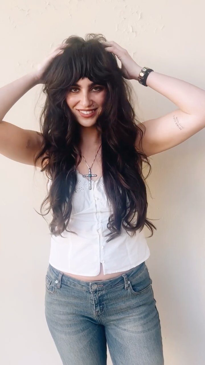 Woman with long dark wavy hair and full bangs posing against a pale wall