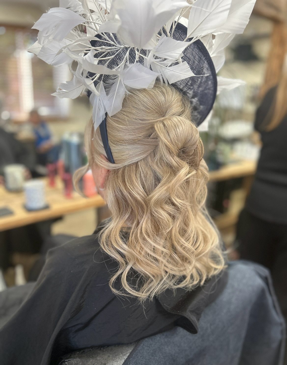 Blonde low updo with loose curls beneath a feathered navy fascinator
