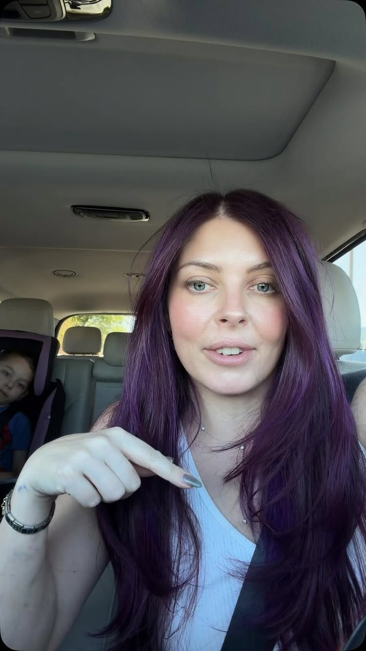 Woman points to her long, vivid dark purple hair while sitting in a car