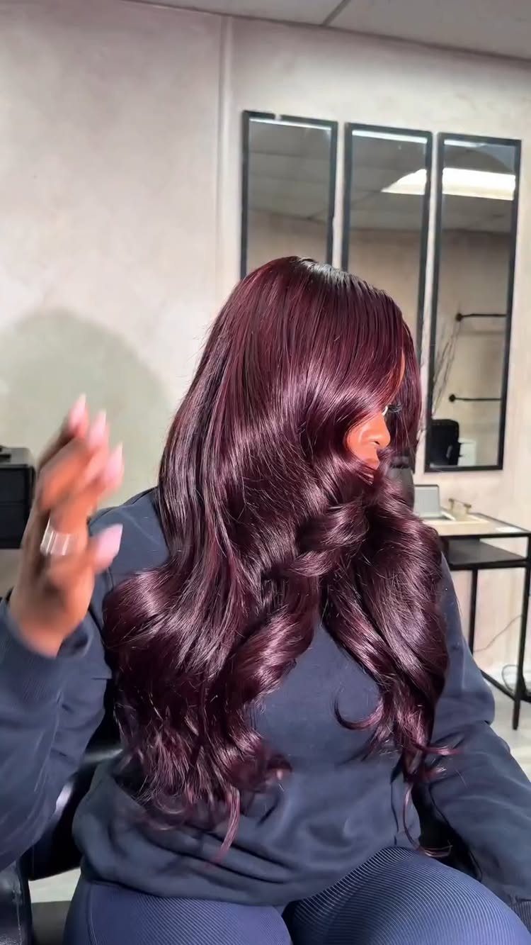 Long burgundy-plum curls fall over a seated person's dark sweatshirt in a salon.