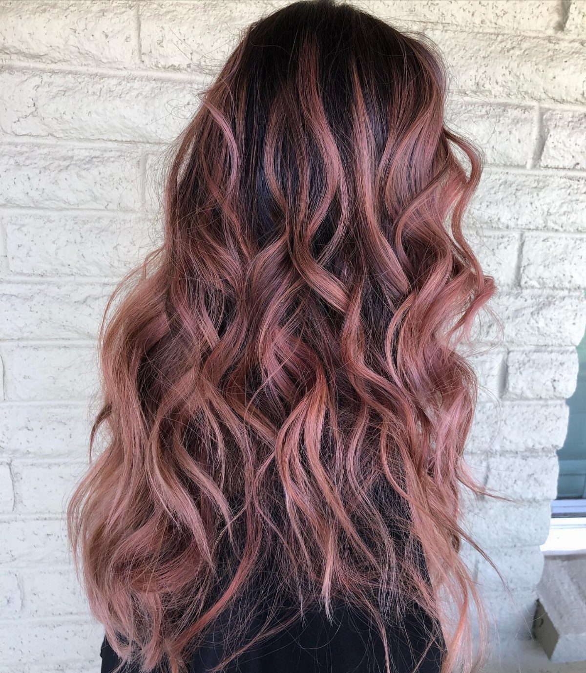 Long dark-rooted hair in muted rose-gold waves against a white brick wall