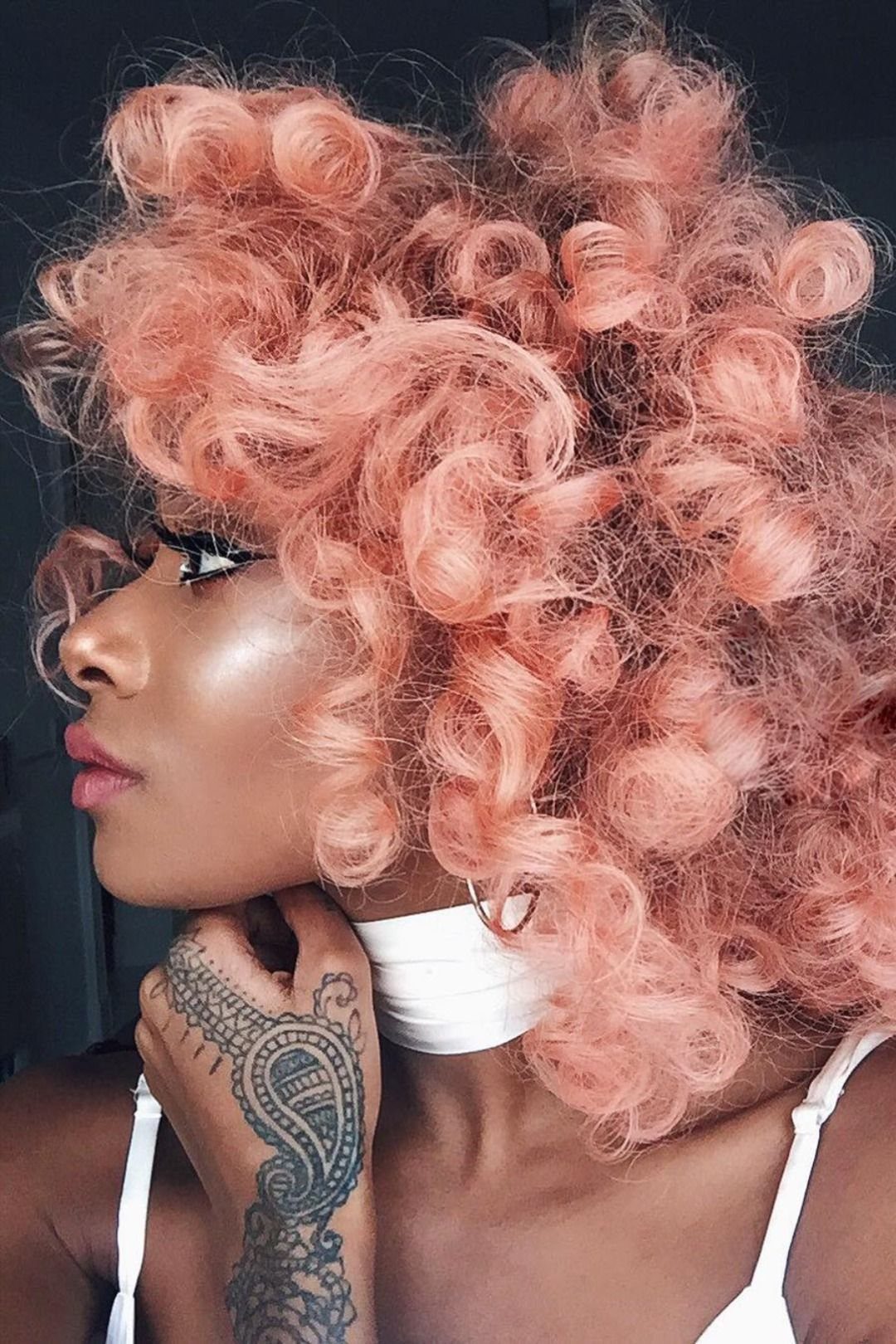 Voluminous peachy-pink curls frame a person’s face against a dark background.