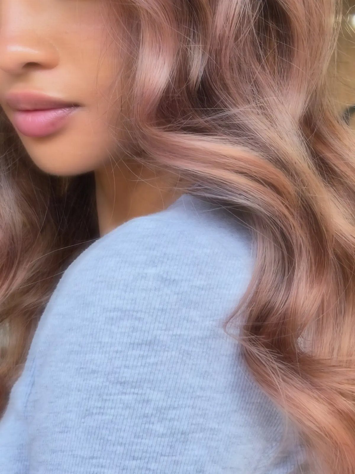 Long rose-gold waves with pink-blonde tones fall over a light blue top