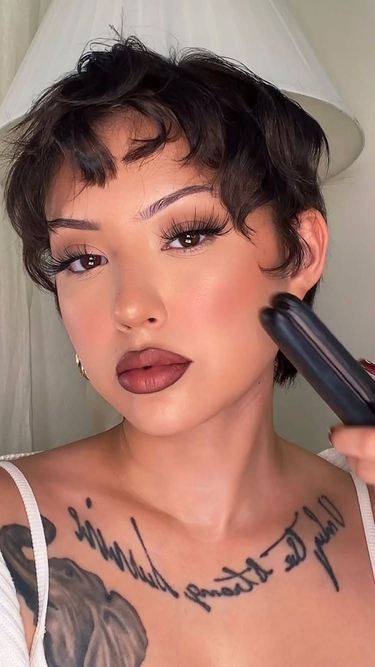 Person with a tousled short black pixie, wispy bangs, makeup, and visible tattoos