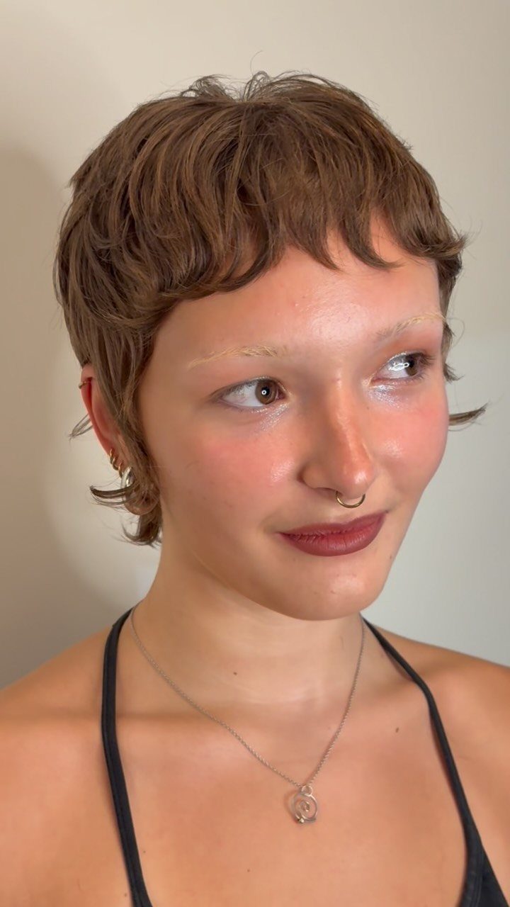 Short tousled brown pixie haircut with bangs on a person posing against a light wall