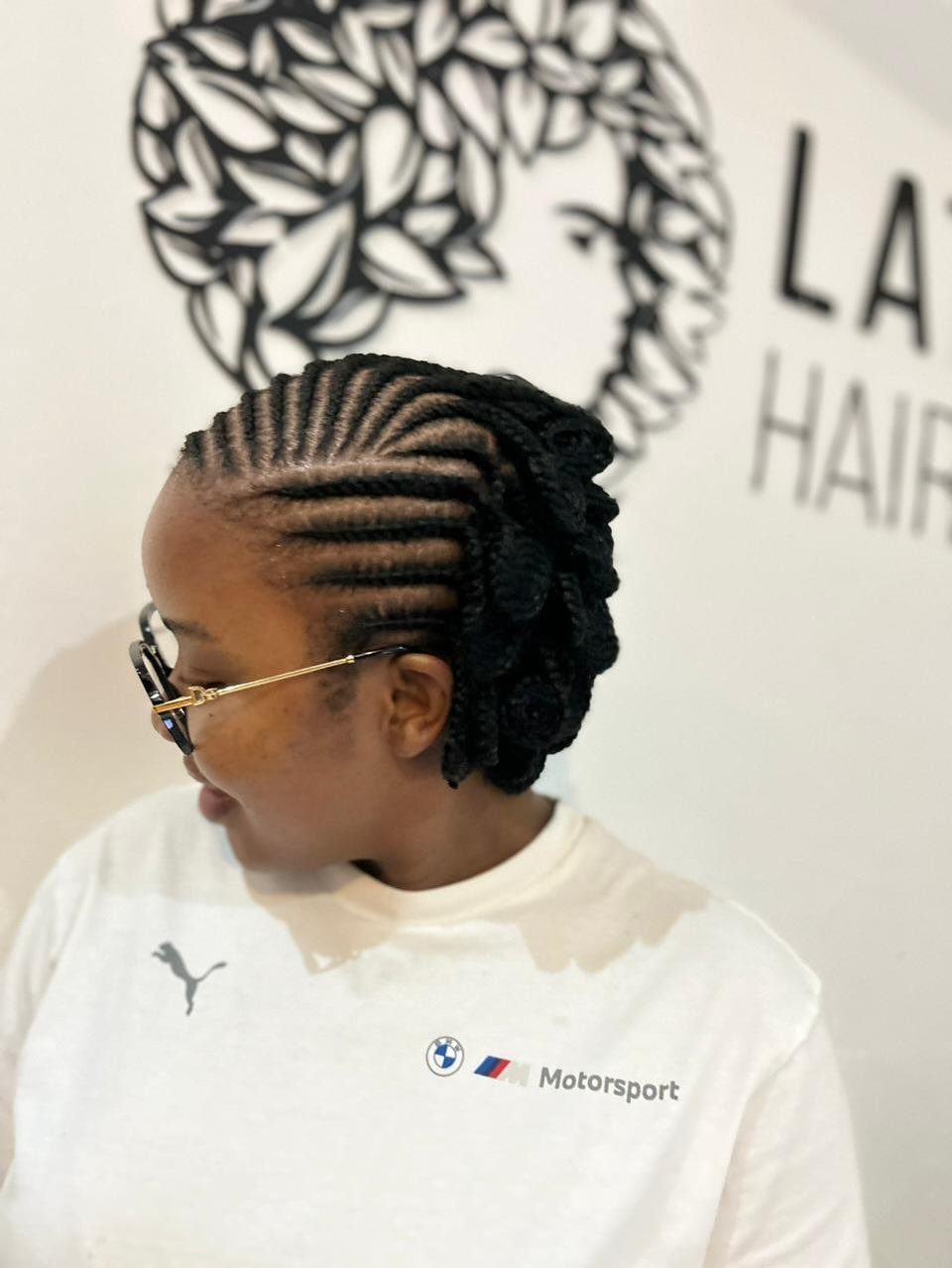 Cornrows swept back into a braided updo on a person wearing glasses