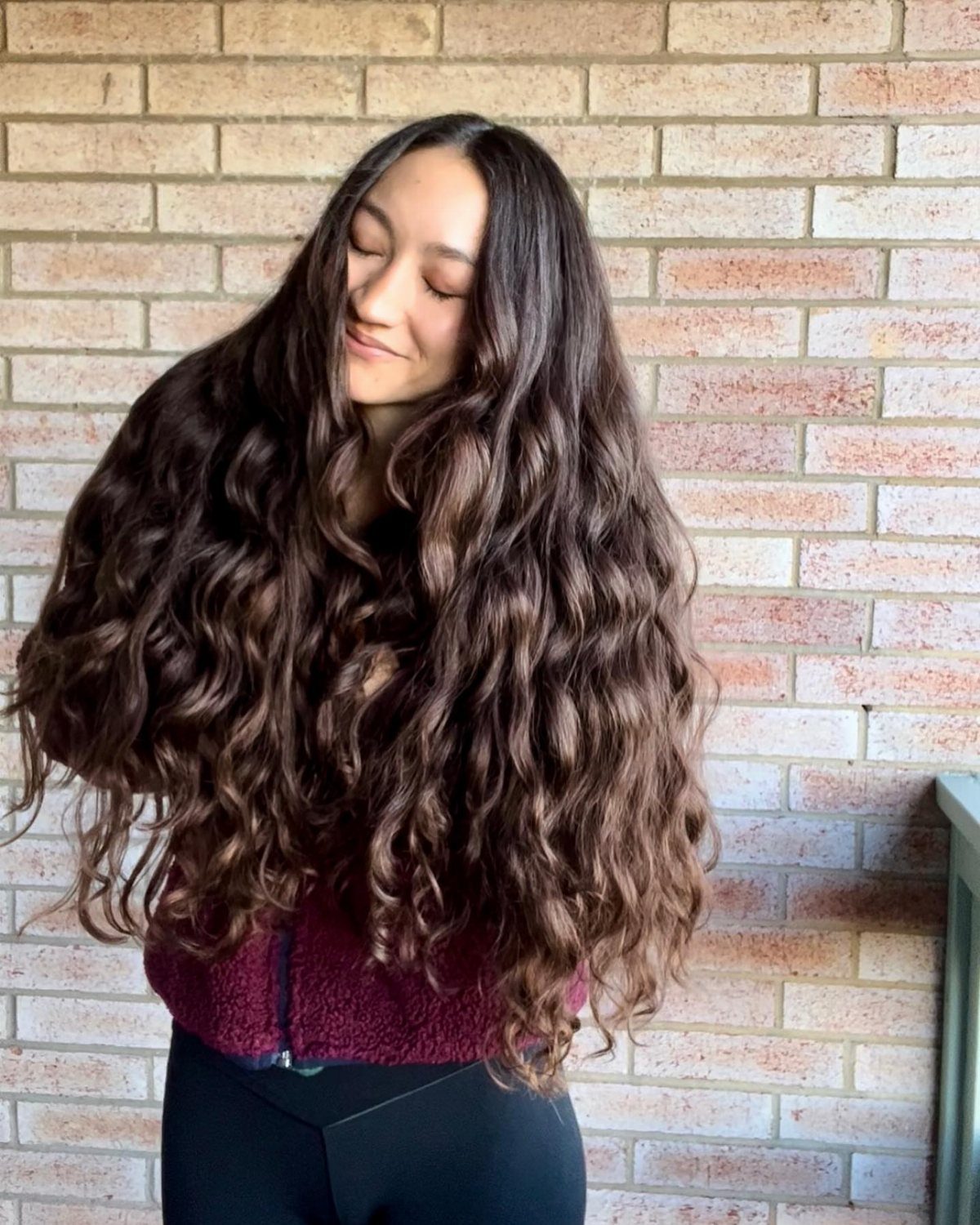 Woman with long, center-parted dark waves standing against a brick wall
